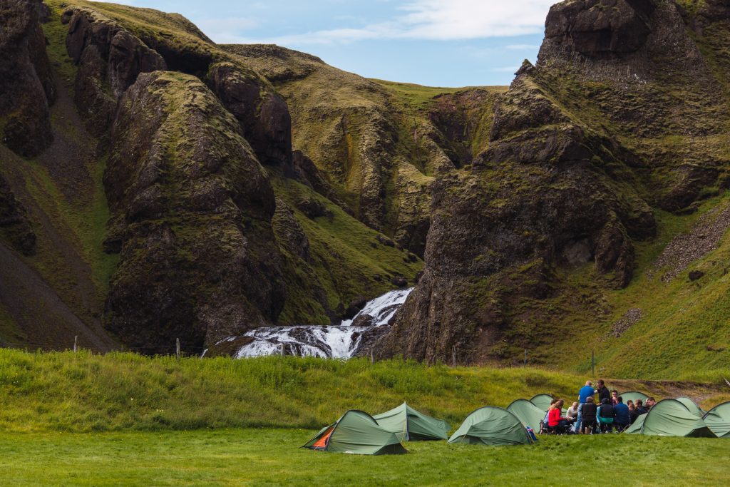 Group of people camping with several green tents in a meadow next to a waterfall in beautiful Iceland